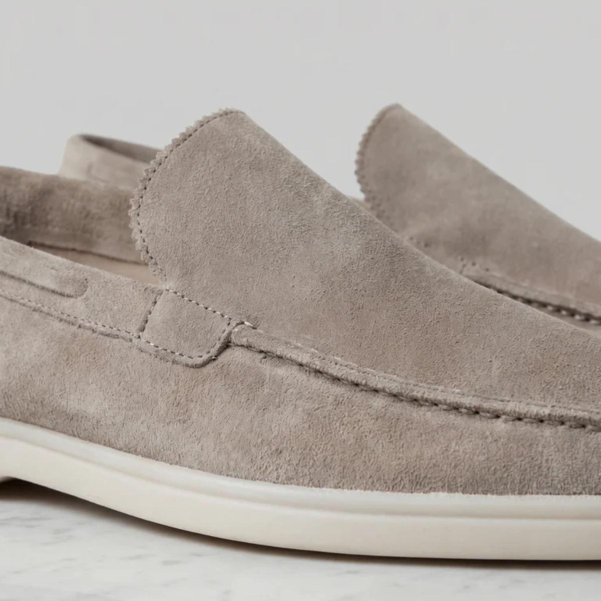 Beige suede slip-on loafer with moc-style paneling, tasseled seam, and white sole
