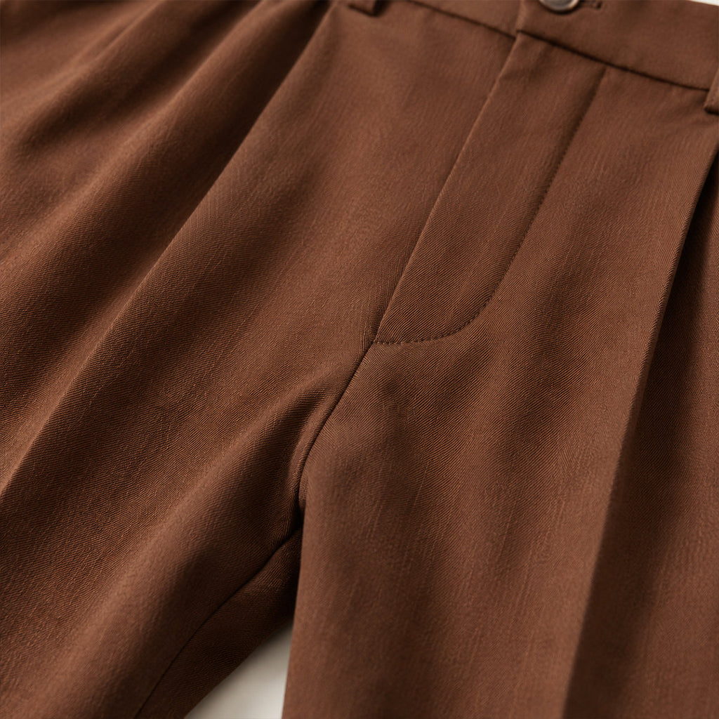 Brown double-pleated pants close-up: front pleats, inset pocket, stitched seam details.